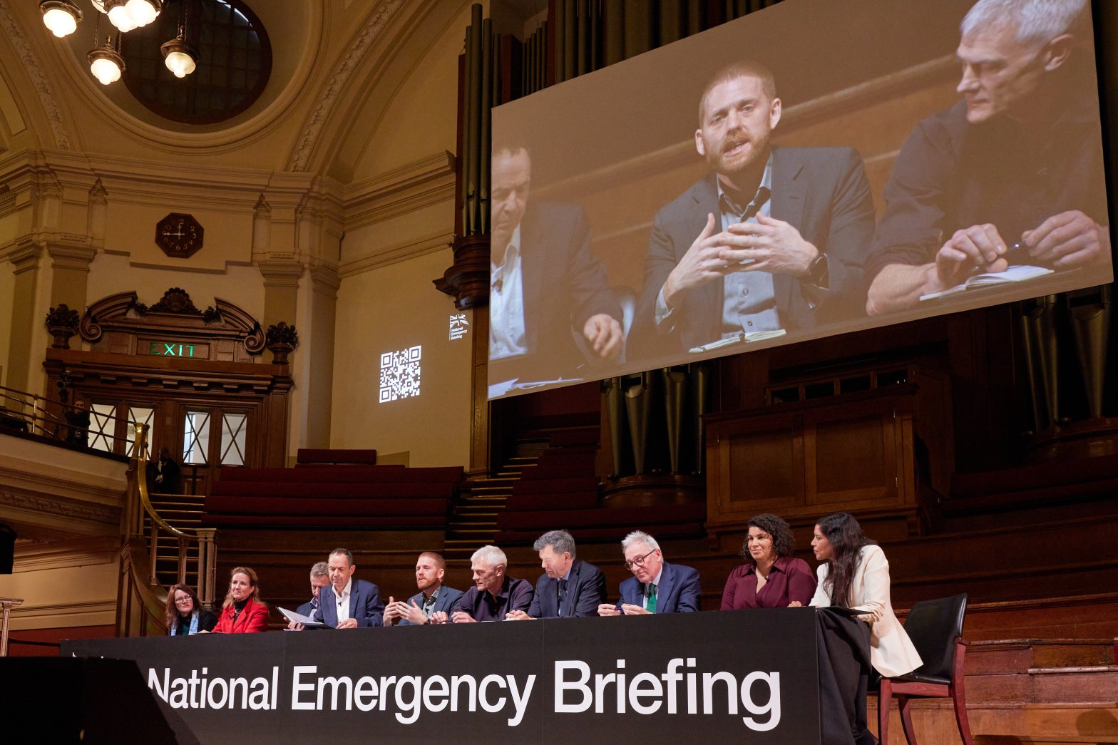 A group pf panellists sat on stage behind a table. Behind them is a screening show a close up of a man talking.